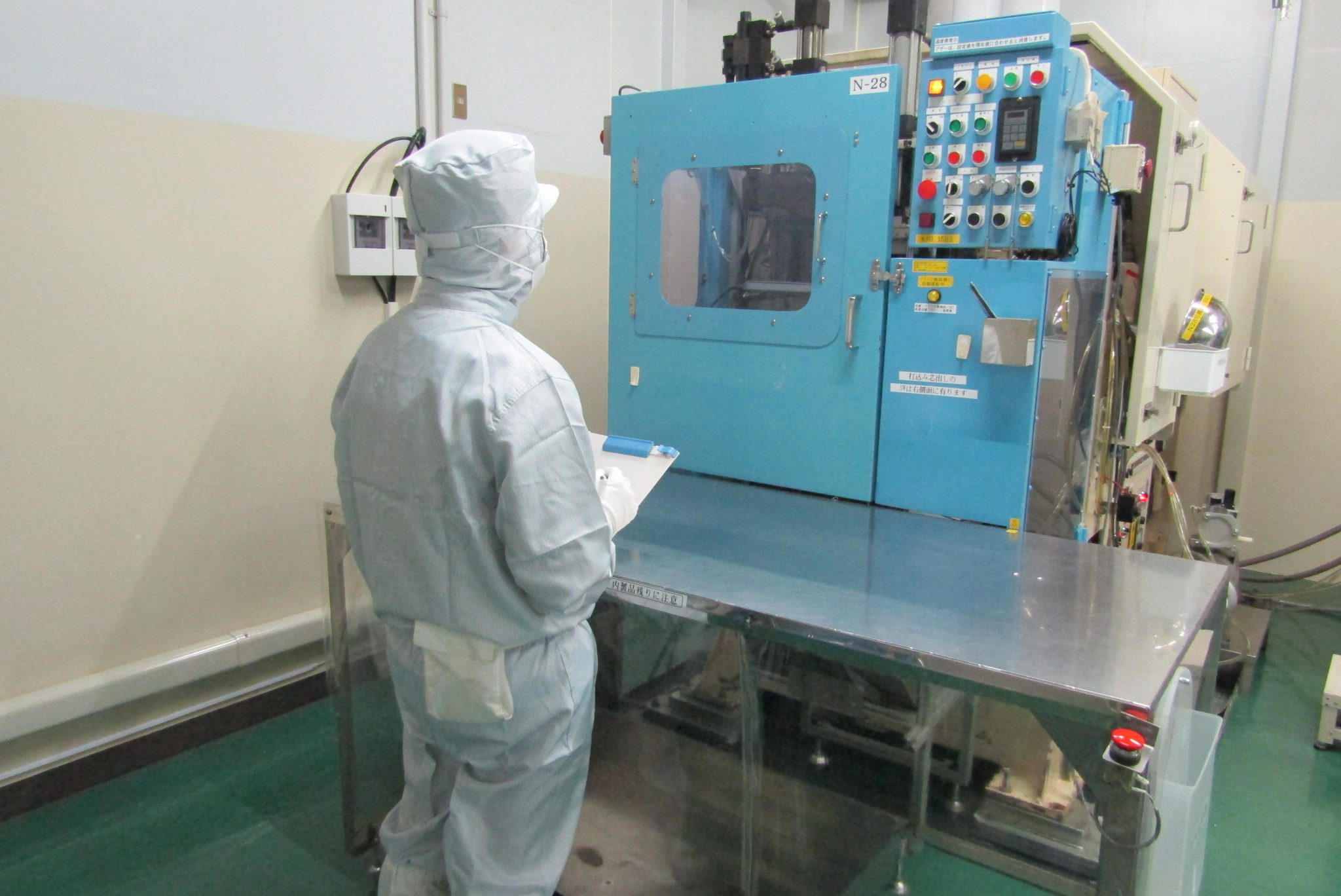 防塵服を着た作業者が、成形機の前で作業記録をつけている